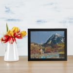 Vintage village at the base of snowy peaks with colorful houses, hills, and a serene river or pond. displayed on the wall