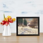 Vintage wall art of a snow-covered pathway with pine trees and a calm winter landscape, framed in black. displayed on the wall