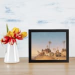Vintage wall art of a desert village with brown huts, palm trees, and a soft blue-beige sky. displayed on the wall