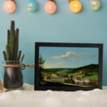 Vintage countryside wall art with hills, houses, and blue sky. displayed on the wall