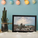 Vintage wall art of a rocky coast with calm blue water and a soft pastel sky in pink, cream, and blue tones. displayed on the wall