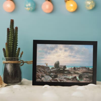 Vintage wall art of a rocky coast with calm blue water and a soft pastel sky in pink, cream, and blue tones. displayed on the wall