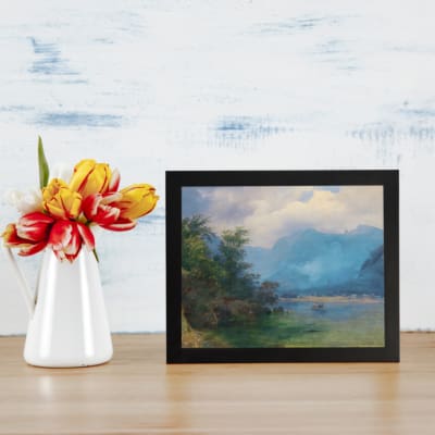 Vintage wall art showcasing mountains, trees, and a water body with blue and green tones, accented by white clouds, framed in a classic design. displayed on the table in a black frame