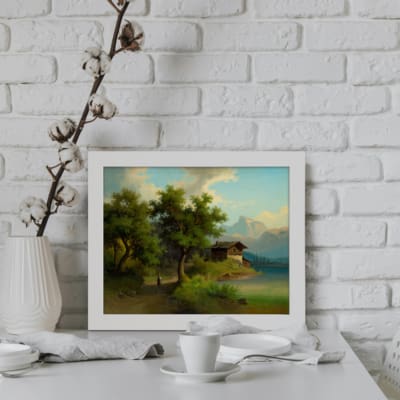Vintage wall art of a rustic countryside landscape with a wooden cabin, lake, trees, and a serene blue sky. displayed on the wall in a white frame
