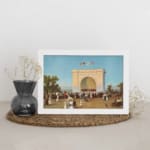 Vintage wall art of a 19th-century crowd in front of a grand white arch with flags, blue sky, and trees, framed in white. displayed on the wall
