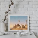 Vintage wall art of a desert village with brown huts, palm trees, and a soft blue-beige sky. displayed on the wall