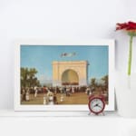 Vintage wall art of a 19th-century crowd in front of a grand white arch with flags, blue sky, and trees, framed in white. displayed on the wall