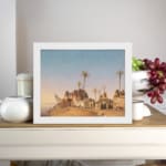 Vintage wall art of a desert village with brown huts, palm trees, and a soft blue-beige sky. displayed on the wall