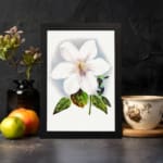 Vintage wall art of a white flower with soft purple hues and green leaves, framed in black. displayed on the wall