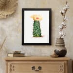 Vintage wall art of a cactus with a yellow flower in bloom, set against a light cream background, framed in black. displayed on the wall