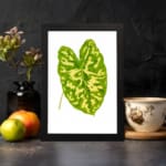 Vintage wall art of a large green tropical leaf with bright yellow streaks on a white background and black frame. displayed on the wall