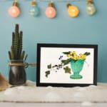 Infidu vintage wall art featuring a blue-green vase with bright yellow flowers, lush green leaves, and delicate purple berries against a light background. displayed on the wall art in a black frame