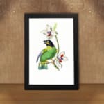 A green bird perched on a branch with white flowers, set on a white background and framed in black. displayed on the wall