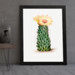 Vintage wall art of a cactus with a yellow flower in bloom, set against a light cream background, framed in black. displayed on the wall