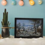 Vintage wall art showing a lively winter village scene with people skating, snowy houses, bare trees, and a soft blue sky. displayed on the wall