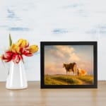 Vintage wall art showing two cows in a grassy field beneath a warm, glowing sky with rolling hills in the background. displayed on the wall