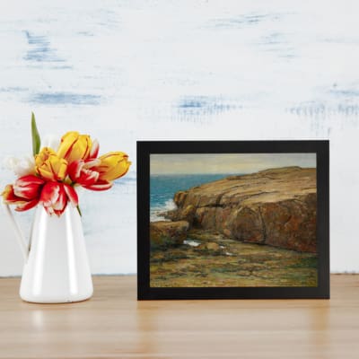 Vintage wall art of a serene rocky seashore with earthy rocks, a calm blue ocean, and a clear sky for coastal or nature-themed decor. displayed on the table in a black frame