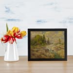 Vintage wall art of a rocky stream with green trees and a cloudy sky in warm earthy tones of brown, green, and yellow. displayed on the wall