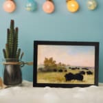 Infidu vintage wall art featuring a calm pastoral scene with black cattle grazing on a green field under a soft, cloudy sky. displayed on the wall art in a black frame