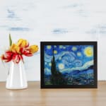 Vincent van Gogh’s The Starry Night vintage wall art shows swirling blue sky, bright yellow stars, a crescent moon, a tree silhouette, and a village. displayed on the wall