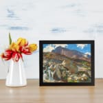 Vintage wall art of a mountain landscape with a waterfall, earthy rocks, green hills, and a blue sky with white clouds. displayed on the wall