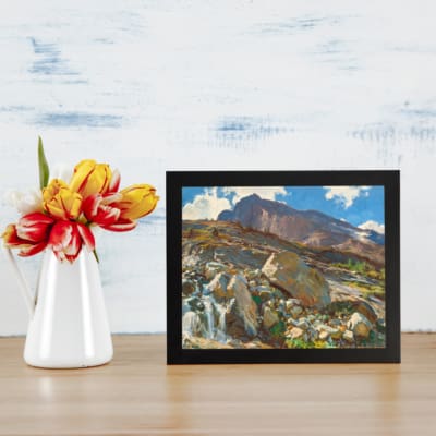 Vintage wall art of a mountain landscape with a waterfall, earthy rocks, green hills, and a blue sky with white clouds. displayed on the wall