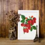 Vintage wall art of red lychee fruits on a branch with green leaves, framed in black on a white background. displayed on the wall