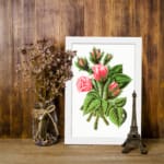 Vintage wall art of soft pink rosebuds with green leaves and stems in a white frame on a white background. displayed on the wall