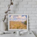 Vintage wall art of a golden grass field under a blue sky with white clouds and trees on the side displayed on the wall