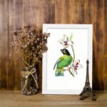 A green bird perched on a branch with white flowers, set on a white background and framed in white. displayed on the wall
