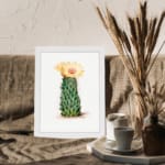 Vintage wall art of a cactus with a yellow flower in bloom, set against a light cream background, framed in white. displayed on the wall