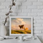 Vintage wall art showing two cows in a grassy field beneath a warm, glowing sky with rolling hills in the background. displayed on the wall