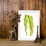 Vintage wall art of a single leaf with flowing, wavy shapes in green and yellow, framed in white on a white background. displayed on the wall