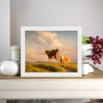Vintage wall art showing two cows in a grassy field beneath a warm, glowing sky with rolling hills in the background. displayed on the wall