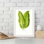 Vintage wall art featuring two large tropical green leaves with white lines and holes, set against a plain white background in a white frame. displayed on the wall