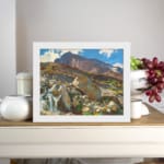 Vintage wall art of a mountain landscape with a waterfall, earthy rocks, green hills, and a blue sky with white clouds. displayed on the wall