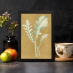 Vintage wall art of pale botanical fronds on an ochre background with a black frame. displayed on the wall in a black frame