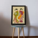 Vintage Japanese wall art with two women in traditional kimonos, featuring rich colors, fine details, and a soft beige background in a black frame. displayed on the table