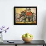 Vintage wall art of a grand elephant parade with red and gold decorations, surrounded by people in a festive village scene. displayed on the wall in a black frame
