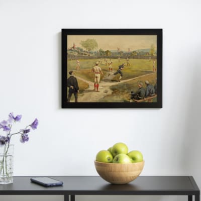 Vintage wall art of a classic baseball game with players in red and blue uniforms on a grassy field, trees and flags in the background. displayed on the wall in a black frame