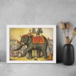 Vintage wall art of a grand elephant parade with red and gold decorations, surrounded by people in a festive village scene. displayed on the wall in a white frame