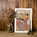 Japanese women in traditional kimonos standing under cherry blossom trees in a serene vintage-style outdoor setting. displayed on the wall in a white frame
