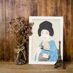 Vintage art of a Japanese woman in a light blue kimono holding a fan with a calm expression and soft background. displayed on the wall in a white frame