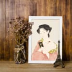 Vintage Japanese wall art featuring a woman in traditional dress holding a book with soft red and green patterns. displayed on the wall in a white frame