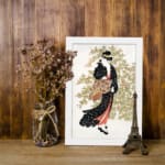 Vintage wall art of a Japanese woman in a black kimono holding a fan, with delicate trees in the background. displayed on the wall in a white frame