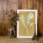 Vintage wall art of pale botanical fronds on an ochre background with a black frame. displayed on the wall in a white frame