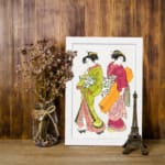 Two Japanese women in traditional green and red kimonos with decorative items, illustrated in elegant vintage style. displayed on the wall in a white frame