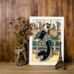 Vintage wall art of a Japanese woman in a black kimono with floral sash, standing before a traditional wooden building. displayed on the wall in a white frame