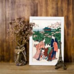 Vintage Japanese wall art of women in kimonos under cherry blossom trees with a temple in the background, displayed on the wall in a white frame