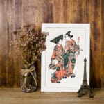 Two figures in traditional Japanese kimonos with orange, green, and black patterns standing gracefully on a plain white background. displayed on the wall in a white frame